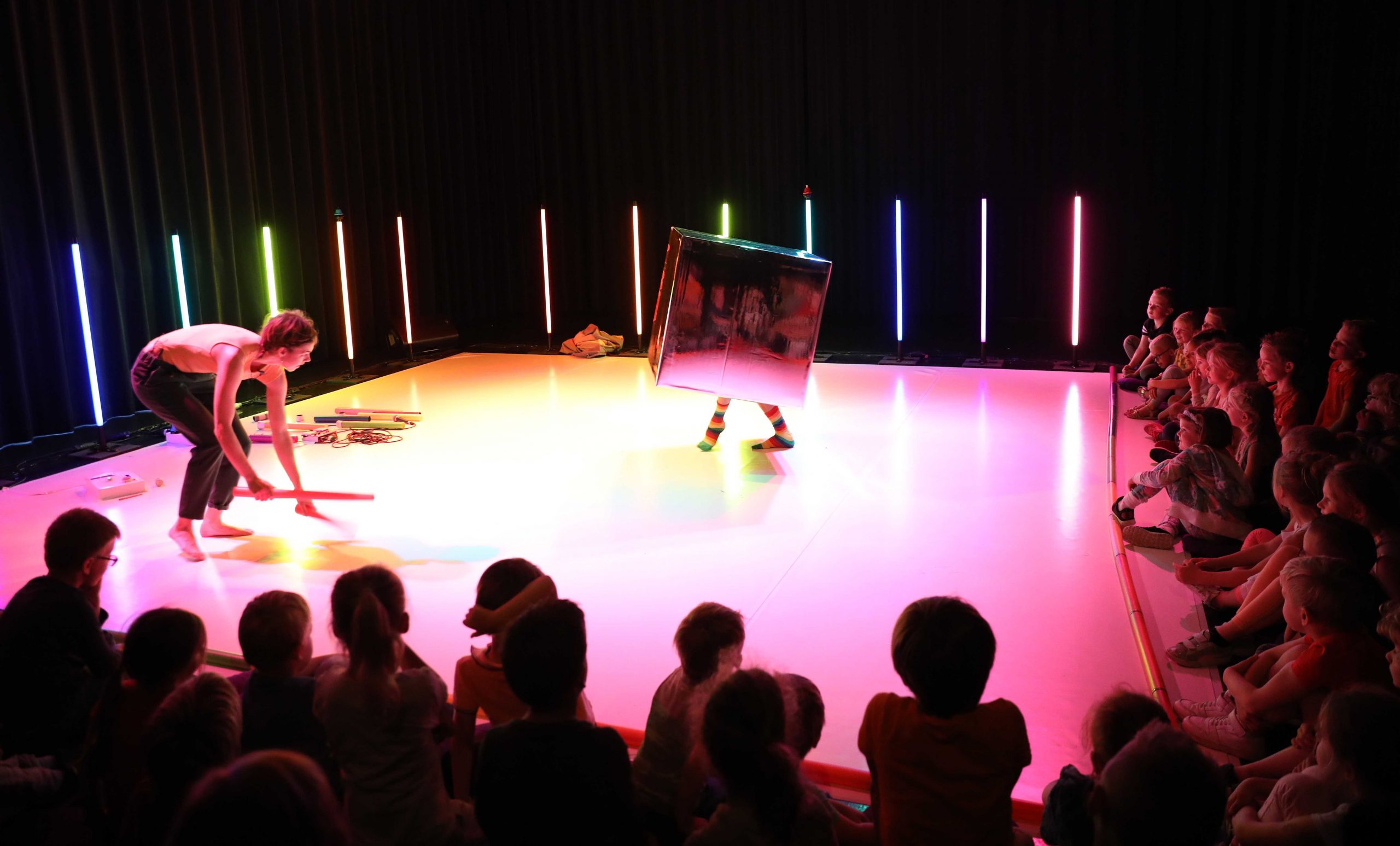 A white stage lit with multi-coloured light, surrounded by glowing multi-coloured sticks. Two figures are the stage; one is crouching down, the other is inside a silver, mirror-like box with only their feet poking through. A young audience sits around two sides of the stage
