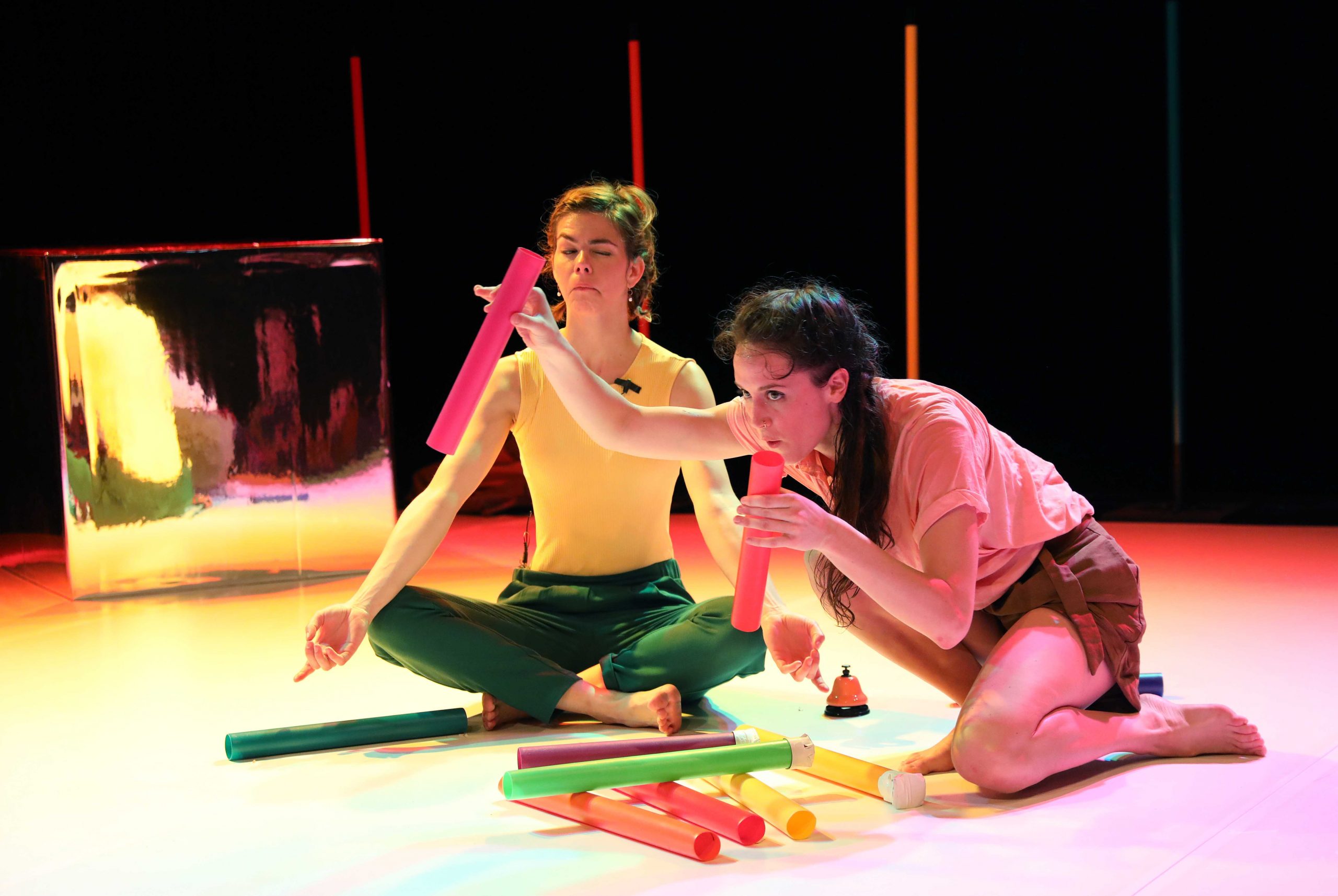 Two figures are on stage. The woman on the left wearing the yellow top and green trousers has her legs crossed. The woman o n the right is holding two pinks tubes and is on one knee. She wears a cream shirt and brown shorts.